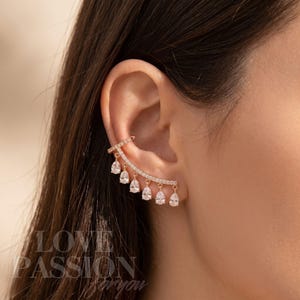 May include: A rose gold ear cuff with a curved bar set with small, clear crystals and six hanging, pear-shaped, clear crystals. The ear cuff is worn on the ear. The text "LOVE PASSION for you" is visible in the lower left corner.