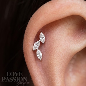 Puede incluir: Una pieza de joyería para piercing de oreja en tono plateado con tres piedras claras de corte marquesa dispuestas en un grupo vertical ligeramente curvado. El texto LOVE PASSION for you es visible en la esquina inferior izquierda.