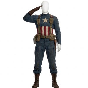 Op de afbeelding: Een mannequin in een Captain America-kostuum. De outfit bestaat uit een blauw shirt met lange mouwen met een witte ster, een rood-wit gestreepte borst en een blauwe broek. Bruine laarzen, een gereedschapsriem en een bruine leren handschoen maken de look compleet.