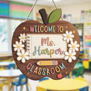 Op de afbeelding: Een houten hangbord in de vorm van een appel met een donkere houtnerf. Het bevat een notitieblokafbeelding met de tekst "WELCOME TO Ms. Harper CLASSROOM", een geel potlood, witte madeliefjes, een groen blad en een klein rood appelicoon.