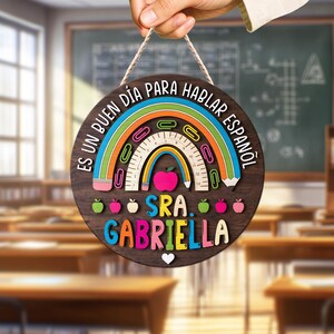 May include: A round wooden sign with a rainbow design and the text "ES UN BUEN D&Iacute;A PARA HABLAR ESPA&Ntilde;OL" in a classroom setting. The sign includes a ruler, paper clips, and an apple. The name "SRA. GABRIELLA" is displayed in colorful letters.