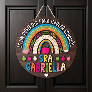 May include: A round wooden sign with a rainbow, apple, and pencil design. The sign has the text "ES UN BUEN DIA PARA HABLAR ESPA&Ntilde;OL" and "SRA. GABRIELLA" in colorful letters. A white heart is at the bottom.