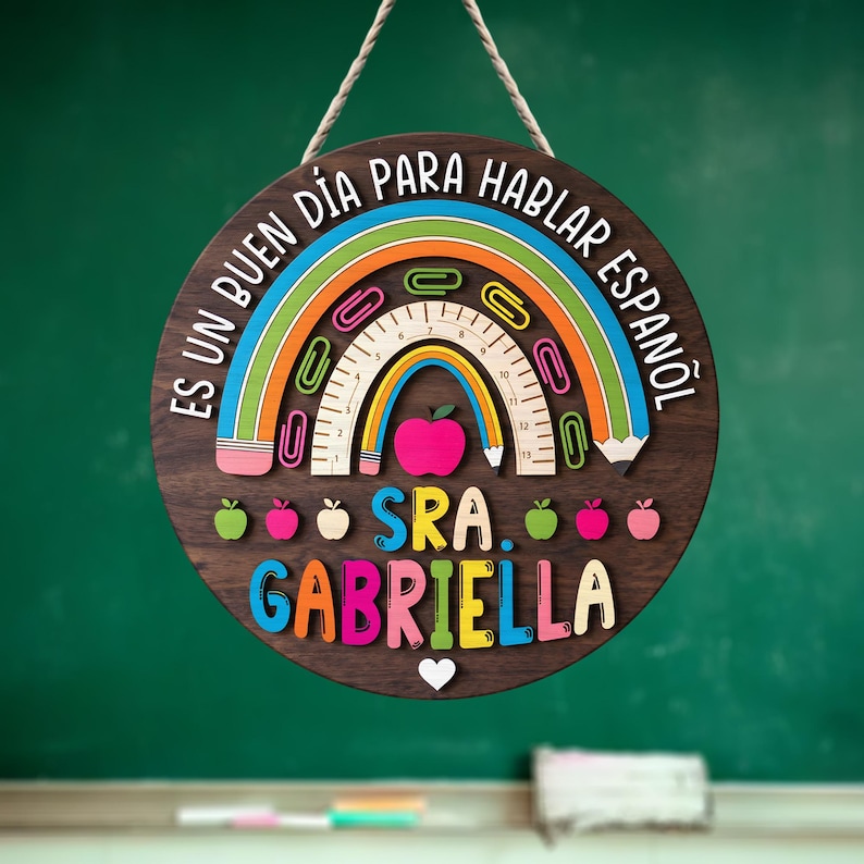 May include: A round wooden sign with a rainbow design and the text "ES UN BUEN D&Iacute;A PARA HABLAR ESPA&Ntilde;OL" in white. The sign also includes the name "SRA. GABRIELLA" in colorful letters, and a heart symbol.
