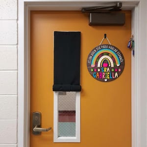 May include: A round wooden sign with a rainbow and apple design, hanging on an orange door. The sign reads "Es un buen dia para hablar espa&ntilde;ol" and "Sra. Gabriella". A black window shade and door handle are also visible.