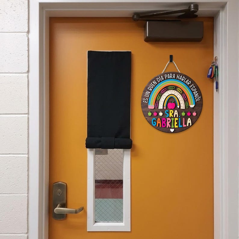 May include: A round wooden sign with a rainbow and apple design, hanging on an orange door. The sign reads "Es un buen dia para hablar espa&ntilde;ol" and "Sra. Gabriella". A black window shade and door handle are also visible.