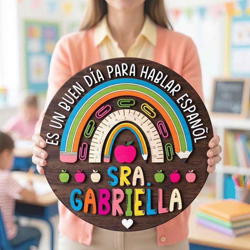 May include: A round wooden sign with a rainbow design and the text "Es un buen d&iacute;a para hablar espa&ntilde;ol" in a classroom setting. The sign includes a ruler, paperclips, and a pink apple. The name "Sra. Gabriella" is displayed in colorful letters.