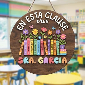 Può includere: Un cartello rotondo in legno con la scritta bianca "EN ESTA CLASE eres". Il cartello presenta fiori colorati, farfalle e una fila di libri con parole positive. Il nome "SRA. GARCIA" è visualizzato in basso.