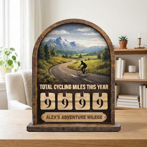 May include: Wooden cycling mileage counter featuring a mountain road scene. The counter shows "9999" miles. Text includes "TOTAL CYCLING MILES THIS YEAR" and "ALEX'S ADVENTURE MILEGE".