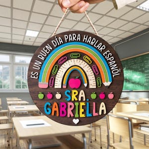 May include: A round wooden sign with a rainbow design, ruler, and apple. The sign has the text "Es un buen d&iacute;a para hablar espa&ntilde;ol" and "Sra. Gabriella" in colorful letters. The sign is held by a hand.