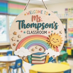 Puede incluir: Un letrero redondo de madera con una cuerda para colgar que incluye el texto "WELCOME TO Ms. Thompson's CLASSROOM". El diseño presenta un arcoíris colorido, un sol amarillo sonriente, nubes blancas, libros apilados, lápices y estrellas.