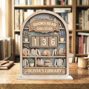 May include: An arched wooden book tracker display featuring the text BOOKS READ THIS YEAR and OLIVIA'S LIBRARY. The shelves contain miniature books, seashells, potted plants, and a three-digit number counter currently set to 136.
