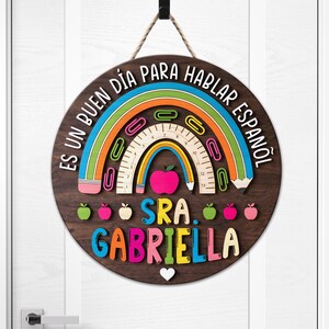 May include: A round wooden sign with a rainbow, apple, and pencil design. The sign has the text "ES UN BUEN D&Iacute;A PARA HABLAR ESPA&Ntilde;OL" and "SRA. GABRIELLA" in colorful letters. A white heart is at the bottom.