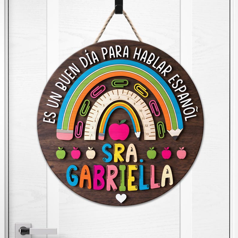 May include: A round wooden sign with a rainbow, apple, and pencil design. The sign has the text "ES UN BUEN D&Iacute;A PARA HABLAR ESPA&Ntilde;OL" and "SRA. GABRIELLA" in colorful letters. A white heart is at the bottom.