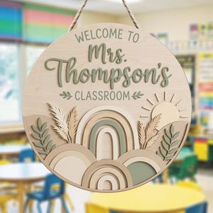 以下が含まれることがあります： ロープ付きの円形木製看板。セージグリーンの文字で「WELCOME TO Mrs. Thompson's CLASSROOM」と書かれています。ベージュ、クリーム、セージグリーンの色合いで、虹、太陽、植物の形にカットされた木材が層状に重ねられたデザインです。