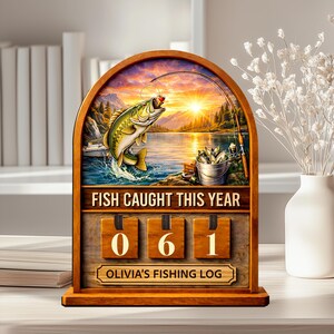 May include: Wooden fishing log with a scenic lake illustration. The sign reads "FISH CAUGHT THIS YEAR" with the number "061" displayed. The bottom of the sign says "OLIVIA'S FISHING LOG". A decorative item for fishing enthusiasts.