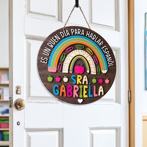 May include: A round wooden sign with a rainbow design and the text "ES UN BUEN D&Iacute;A PARA HABLAR ESPA&Ntilde;OL" and "SRA. GABRIELLA". The sign is hanging on a white door. The rainbow includes a ruler, paperclips, and a pink apple.