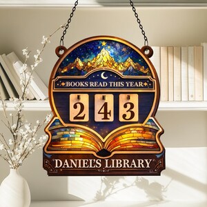 Puede incluir: Un letrero de madera con un diseño celestial y el texto "BOOKS READ THIS YEAR". Cuenta con un contador que muestra "243" y las palabras "DANIEL'S LIBRARY". El letrero está colgado de una cadena.