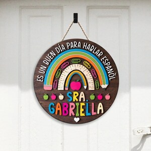 May include: A round wooden sign with the text "ES UN BUEN D&Iacute;A PARA HABLAR ESPA&Ntilde;OL" and "SRA. GABRIELLA". The sign features a rainbow, apple, pencil, and paperclip design. It is hanging from a black hook with a rope.