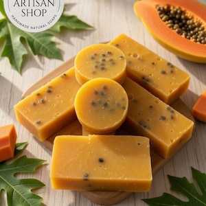 May include: Handmade soaps in yellow-orange hues, some rectangular, some circular, with dark specks. The soaps are arranged on a wooden board, alongside papaya slices and leaves. The shop name is "Lena's Artisan Shop".
