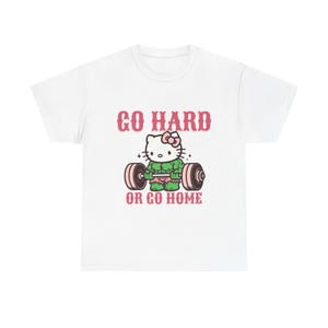 以下が含まれることがあります： 白いTシャツ。漫画のハローキティのキャラクターがバーベルを持ち上げています。キャラクターは緑色で筋肉質で、ピンクのリボンとショーツをしています。「GO HARD」と「OR GO HOME」の文字がキャラクターの上と下にピンク色で書かれています。