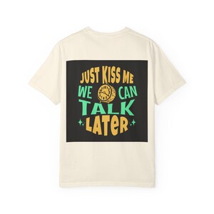 May include: Cream-colored t-shirt with a black square graphic. The graphic features the text "JUST KISS ME WE CAN TALK LATER" in yellow, green, and orange, with a clock illustration. The shirt is made of a soft material.