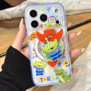 Peut inclure: Coque de téléphone transparente avec des personnages de Toy Story. La coque a une poignée pop-up avec un design d'alien vert et le mot "PRIZE". D'autres personnages incluent Woody, Buzz Lightyear et un dinosaure vert. Le mot "STORY" est également visible.