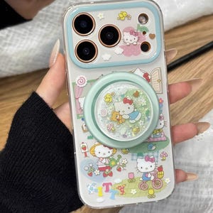 May include: Clear phone case with a light blue border and a pop socket. The case features a colorful design with Hello Kitty characters, flowers, and the text "T". The pop socket has a glittery, snow globe effect.