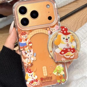May include: Clear phone case with a gold-colored back and a cartoon design featuring Duffy and Friends characters. The case has a pop-up grip with a cartoon dog character wearing a red hat. The case is decorated with various cartoon characters.