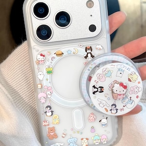 May include: Clear phone case with colorful cartoon character stickers, including Hello Kitty, penguins, and bears. A matching pop socket is attached. The case has a silver camera section and the text "CCxCase" is visible.