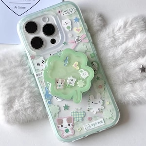 Op de afbeelding: Transparante telefoonhoes met een lichtgroene rand, versierd met cartoonstickers van dieren, sterren en harten. Een groene klavervormige telefoonhouder is bevestigd. De hoes heeft de tekst "CCxCase" onderaan.