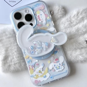 May include: Clear phone case with a cartoon design featuring a white bunny, clouds, and stars. The case has a pop-up grip in the shape of the bunny. The text "Dream" is visible on the case. The case is decorated with blue, yellow, and pink accents.