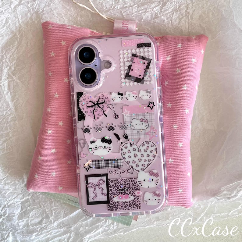 Op de afbeelding: Een roze telefoonhoesje met een doorzichtige rand, versierd met Hello Kitty-ontwerpen, harten, strikken en andere schattige afbeeldingen. De hoes ligt op een roze kussen met witte sterpatronen.