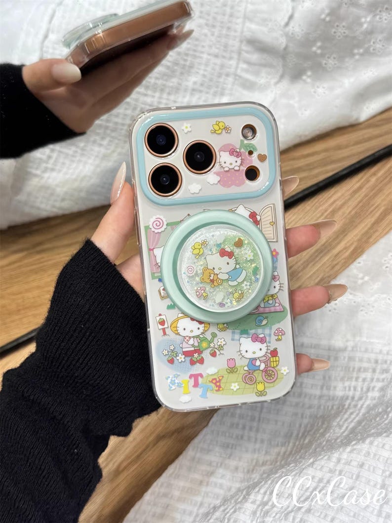 May include: Clear phone case with a light blue border and a pop socket. The case features a colorful design with Hello Kitty characters, flowers, and the text "T". The pop socket has a glittery, snow globe effect.