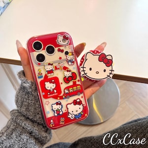 May include: A red and clear phone case featuring Hello Kitty graphics and a matching pop socket. The case has a red border and is decorated with various Hello Kitty illustrations, including the character on a bus and holding a strawberry.