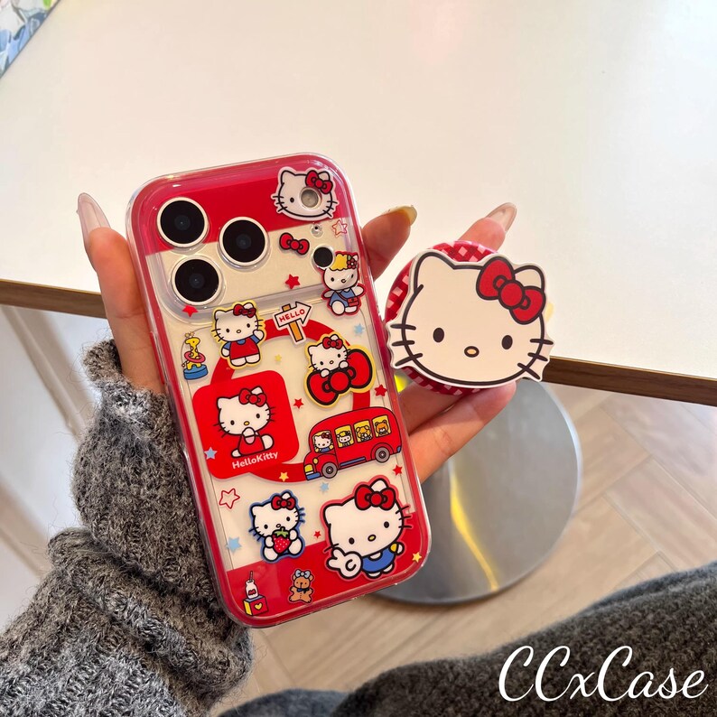 May include: A red and clear phone case featuring Hello Kitty graphics and a matching pop socket. The case has a red border and is decorated with various Hello Kitty illustrations, including the character on a bus and holding a strawberry.