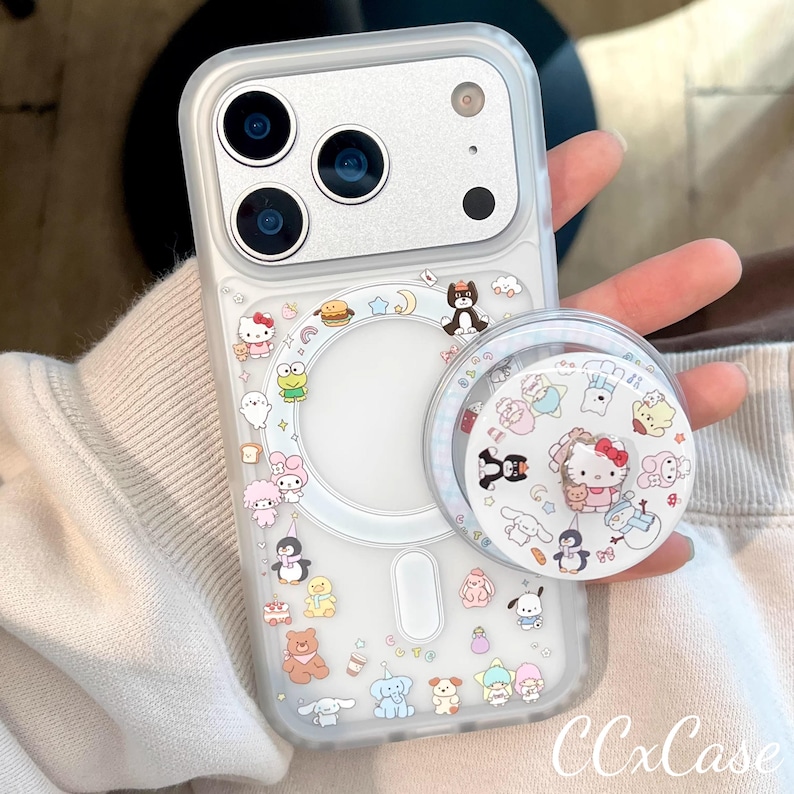 May include: Clear phone case with colorful cartoon character stickers, including Hello Kitty, penguins, and bears. A matching pop socket is attached. The case has a silver camera section and the text "CCxCase" is visible.