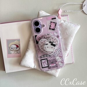 Op de afbeelding: Een doorzichtige telefoonhoes met een roze achtergrond met Hello Kitty-ontwerpen, harten en een pop-up grip. De hoes heeft een hartvormige pop-up grip met een Hello Kitty gezicht. De hoes ligt op een witte ondergrond met een roze boek.