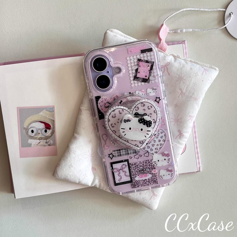 Op de afbeelding: Een doorzichtige telefoonhoes met een roze achtergrond met Hello Kitty-ontwerpen, harten en een pop-up grip. De hoes heeft een hartvormige pop-up grip met een Hello Kitty gezicht. De hoes ligt op een witte ondergrond met een roze boek.