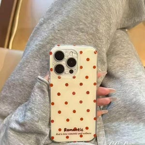 Puede incluir: Una funda transparente para tel&eacute;fono con fondo color crema y un patr&oacute;n de lunares rojos espaciados. La funda tiene un recorte para la c&aacute;mara y la palabra "Romantic" con el texto "Red is love romantic and wellness" impreso en la parte inferior.