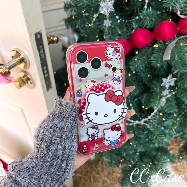 May include: A clear phone case with a red border and a pop-up grip featuring the Hello Kitty character. The case has a red and white checkered pattern and multiple Hello Kitty illustrations. The case is designed to fit a phone with three camera lenses.
