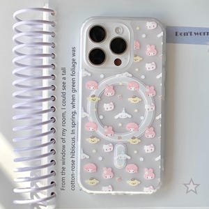 May include: Clear phone case with a pattern of pink and white cartoon characters, stars, and clouds. The case has a circular cutout for wireless charging. Text on the case reads "CCxCase". The case is on a white surface with text from a notebook.