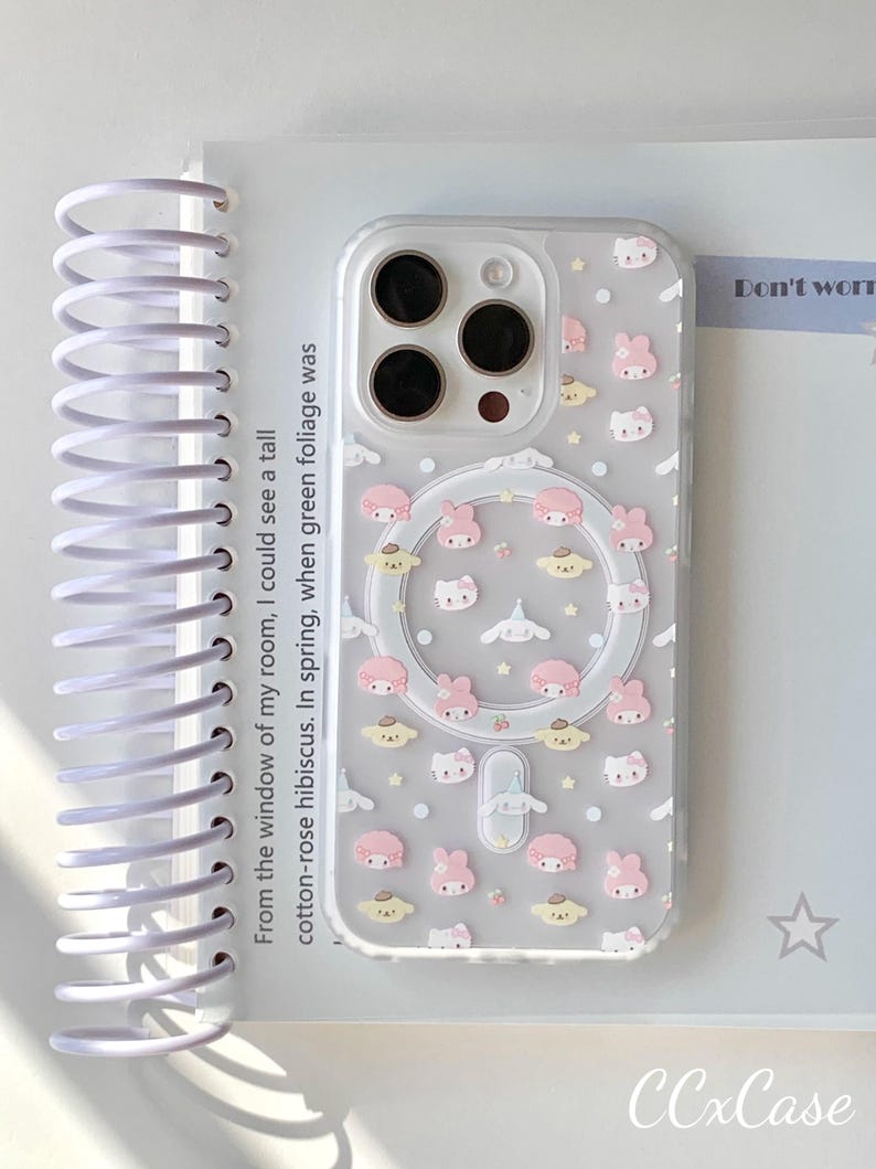 May include: Clear phone case with a pattern of pink and white cartoon characters, stars, and clouds. The case has a circular cutout for wireless charging. Text on the case reads "CCxCase". The case is on a white surface with text from a notebook.