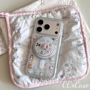 May include: Clear phone case with a pop socket featuring cartoon characters. The case is decorated with small, colorful figures. The phone case rests on a white and pink patterned fabric with a pink border. The text "CCxCase" is visible.