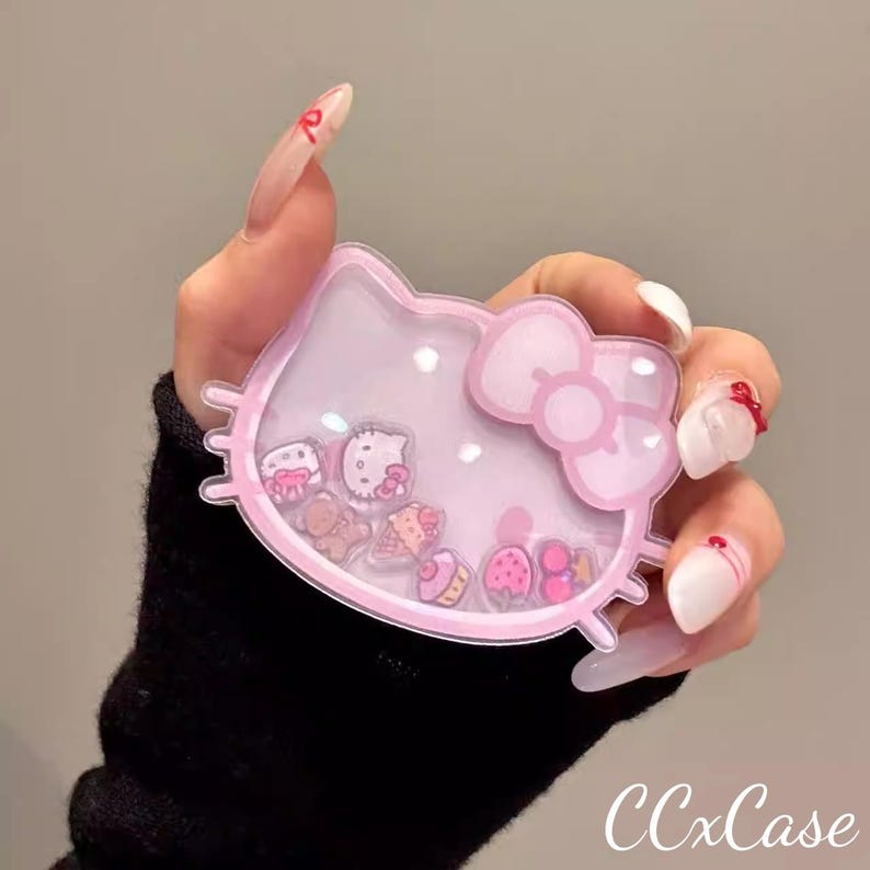 May include: A pink, Hello Kitty-shaped phone case with a clear, liquid-filled center containing small, floating charms. The case has a bow detail and the text "CCxCase" in the bottom right corner.
