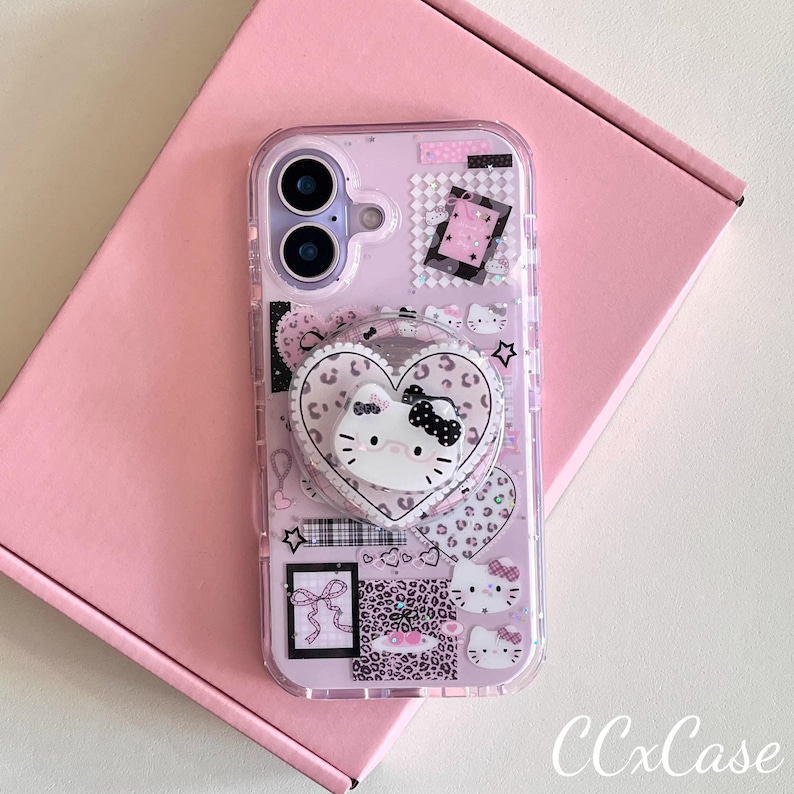 Op de afbeelding: Transparante telefoonhoes met een roze achtergrond, met een hartvormige popsocket met een Hello Kitty-ontwerp. De hoes heeft een collage van roze en zwarte patronen, waaronder luipaardprint en kleine Hello Kitty-gezichten. De tekst "CCxCase" is zichtbaar.