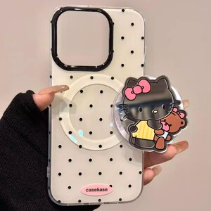 May include: Clear phone case with black polka dots and a black border around the camera cutout. A black Hello Kitty pop socket with a pink bow and a teddy bear is attached. The text "casekase" is visible.