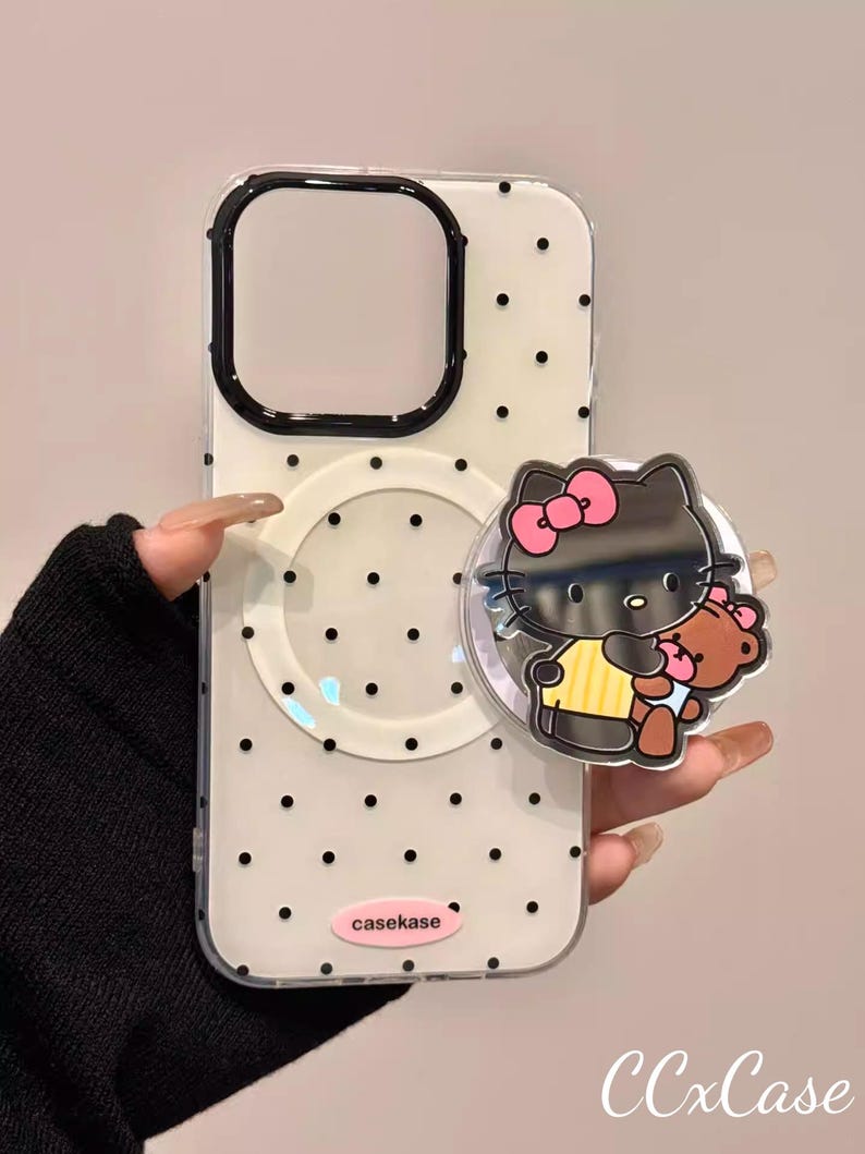 May include: Clear phone case with black polka dots and a black border around the camera cutout. A black Hello Kitty pop socket with a pink bow and a teddy bear is attached. The text "casekase" is visible.