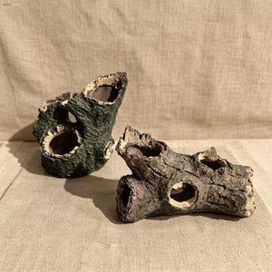 Puede incluir: Dos adornos decorativos para acuario con forma de troncos huecos. Los troncos artificiales están pintados en tonos verdes, marrones y beige, con aberturas de varios tamaños. La superficie texturizada imita la apariencia de la corteza de los árboles, añadiendo un toque natural al acuario.