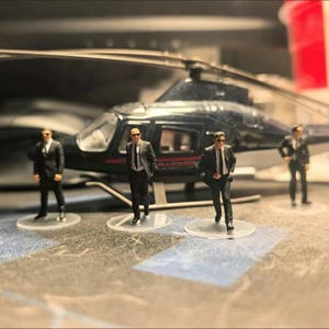 May include: Miniature black helicopter with a silver rotor and four small figurines of people in black suits and sunglasses. The figurines are standing on clear circular bases. The background is blurred.