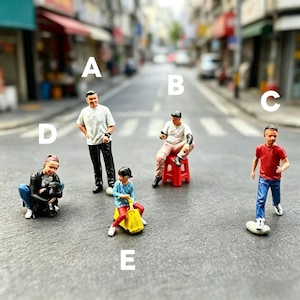 May include: A collection of miniature figurines depicting people in different poses, such as standing, sitting on a red stool, and a child riding a yellow toy. The figures are positioned on a street, with a blurred background of buildings and a road.